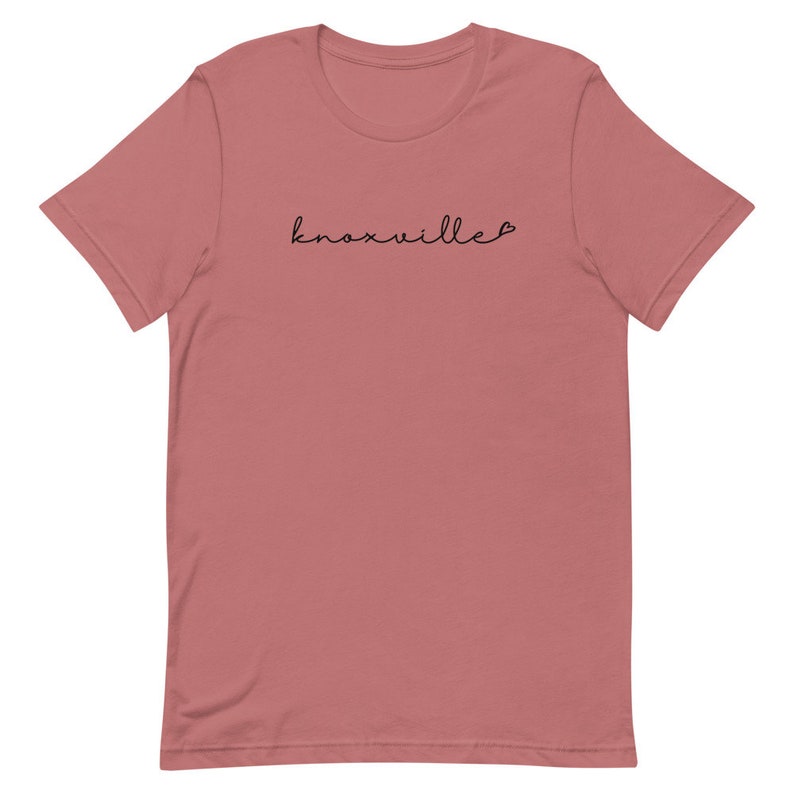 May include: A dusty rose t-shirt with the word "Knoxville" in a delicate, black script across the chest. The script has a small heart at the end of the word. The shirt has short sleeves and a crew neck.