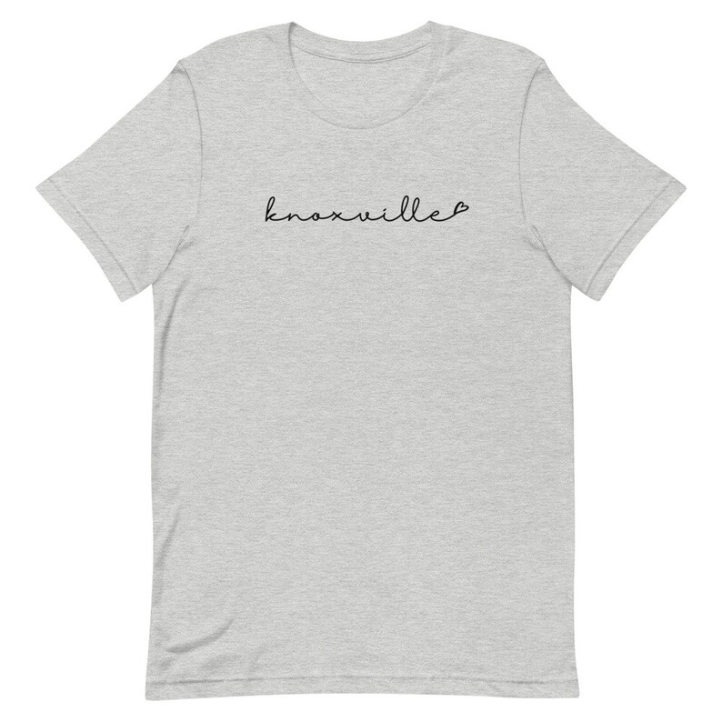 May include: A light gray t-shirt with the word "Knoxville" in a cursive font. The text is in black. The t-shirt has short sleeves and a crew neck.