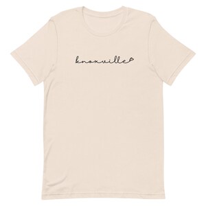 May include: A cream-colored t-shirt with the word "Knoxville" in a black cursive font. A small heart follows the word. The shirt has short sleeves and a crew neck.