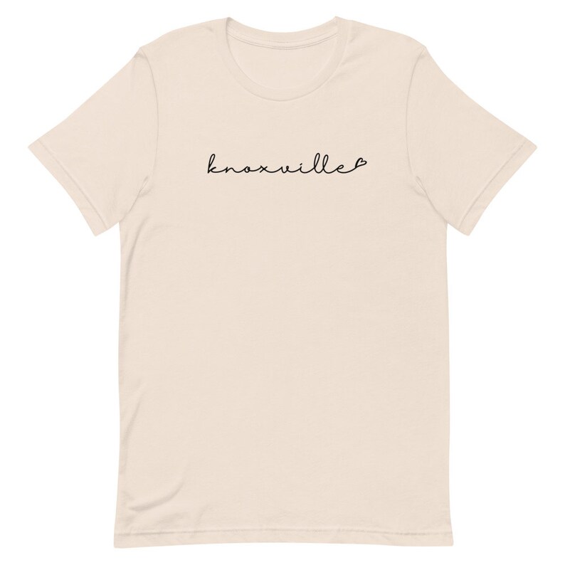 May include: A cream-colored t-shirt with the word "Knoxville" in a black cursive font. A small heart follows the word. The shirt has short sleeves and a crew neck.