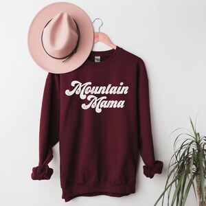 May include: Burgundy sweatshirt with white text that reads "Mountain Mama" in a retro font. The sweatshirt is hanging on a wooden hanger against a white wall.