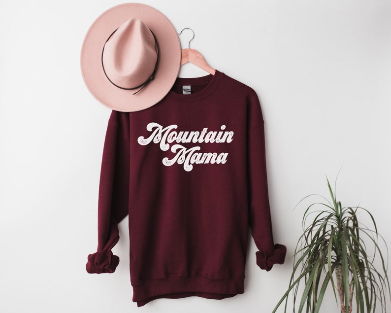 May include: Burgundy sweatshirt with white text that reads "Mountain Mama" in a retro font. The sweatshirt is hanging on a wooden hanger against a white wall.