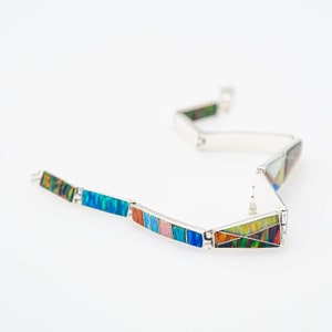 May include: A silver bracelet with rectangular and triangular segments. Each segment features a mosaic of colorful, iridescent stones in shades of blue, green, red, and yellow. The bracelet is displayed on a white background.