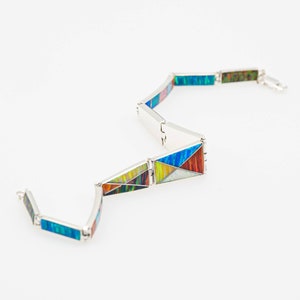 May include: A silver bracelet with geometric inlays of various colors, including blue, red, yellow, and green. The bracelet is composed of rectangular links connected by hinges, creating a flexible design. The inlays have a mosaic appearance.