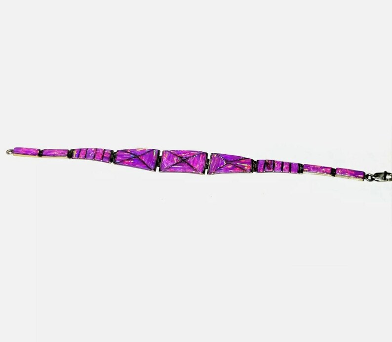 Pode incluir: Uma pulseira roxa com formas geom&eacute;tricas. A pulseira apresenta elos retangulares e trapezoidais em tons de roxo, conectados por elos pretos. O fecho &eacute; prateado. A pulseira tem aproximadamente 18 cm de comprimento.