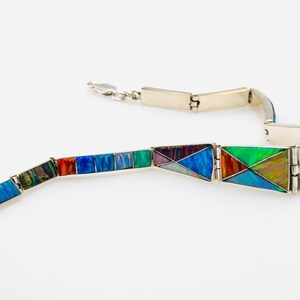 May include: A silver bracelet with a hinged design features colorful opal inlay. The bracelet is made up of multiple sections that are connected by hinges. The opal inlay is a mix of blues, greens, reds, and purples.