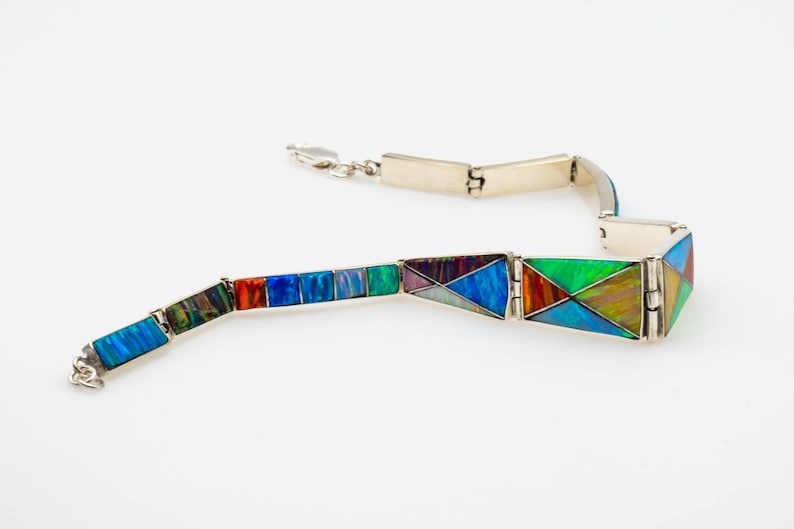 May include: A silver bracelet with a hinged design features colorful opal inlay. The bracelet is made up of multiple sections that are connected by hinges. The opal inlay is a mix of blues, greens, reds, and purples.