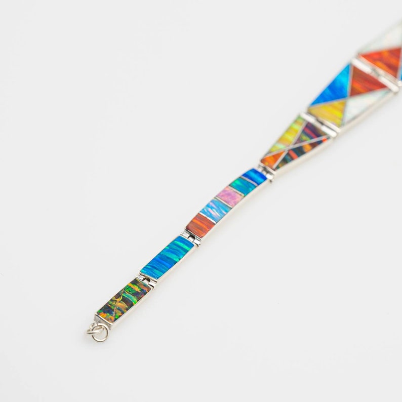May include: A silver bracelet with colorful geometric opal inlays. The bracelet features a variety of shapes, including triangles and rectangles, in shades of blue, orange, yellow, and pink. The bracelet is set against a white background.