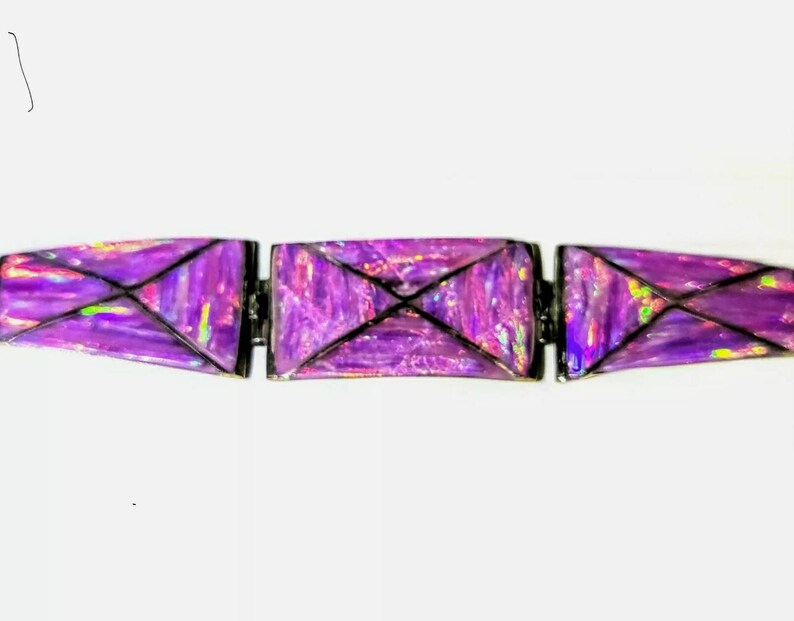 Pode incluir: Uma vista de perto de uma pulseira roxa com um design geom&eacute;trico. A pulseira &eacute; composta por tr&ecirc;s segmentos retangulares ligados por dobradi&ccedil;as. Cada segmento apresenta uma cor roxa vibrante e iridescente com detalhes em preto, criando um efeito visual marcante. A pulseira est&aacute; sobre um fundo branco.