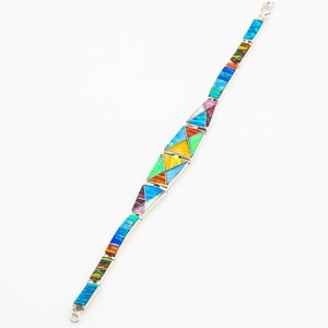 May include: A silver bracelet with a geometric design made of colorful inlaid stones. The stones are arranged in a pattern of triangles and rectangles. The bracelet is made of sterling silver and has a clasp closure.