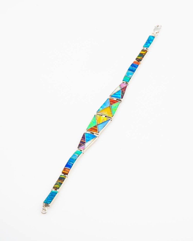 May include: A silver bracelet with a geometric design made of colorful inlaid stones. The stones are arranged in a pattern of triangles and rectangles. The bracelet is made of sterling silver and has a clasp closure.