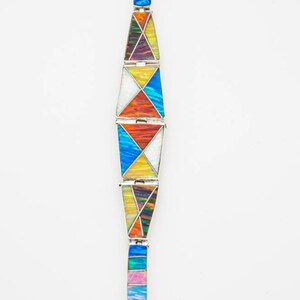 May include: A silver bracelet with geometric shapes in various colors, including blue, orange, yellow, and white. The bracelet is made of multiple connected segments, each with a unique pattern. The background is plain white.