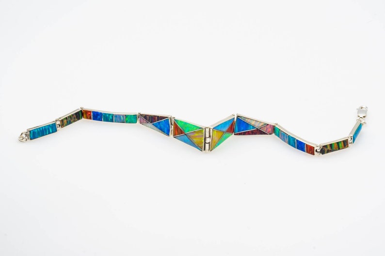 May include: A colorful bracelet made of inlaid stone and silver. The bracelet is made up of rectangular and triangular pieces of stone, each with a different pattern and color. The bracelet is open and has a clasp.