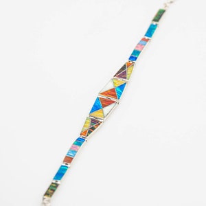 May include: A silver bracelet with colorful geometric designs. The bracelet features a series of rectangular and triangular segments in various colors, including blue, orange, yellow, and pink. The bracelet is set against a white background.