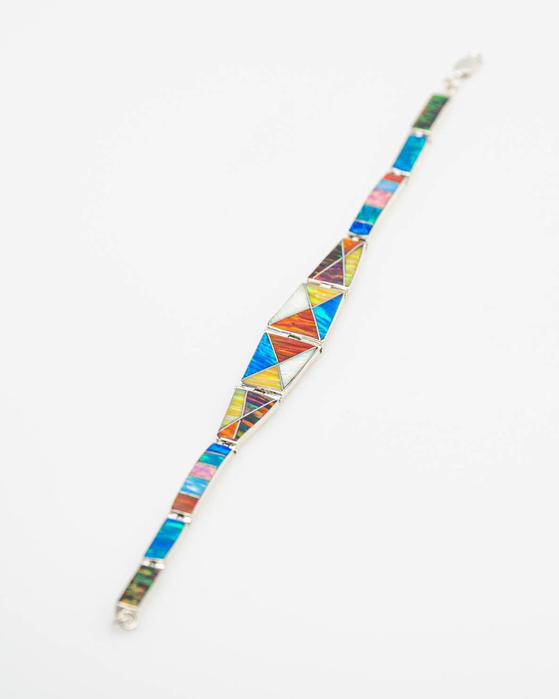 May include: A silver bracelet with colorful geometric designs. The bracelet features a series of rectangular and triangular segments in various colors, including blue, orange, yellow, and pink. The bracelet is set against a white background.