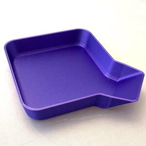 May include: A solid purple tray with a unique shape. The tray has a rectangular base with a cut-out on one side, and a raised edge. The surface has a textured appearance.