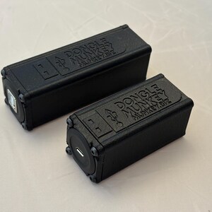 May include: Two black rectangular devices with rounded corners are displayed. The devices have the text "DONGLE MUNKEY MUNKEY7.BIZ" embossed on the top. One device is larger than the other. The smaller device has a USB port.