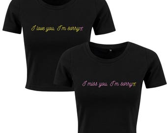 Puede incluir: Dos camisetas negras recortadas de manga corta. La camiseta de arriba tiene el texto "I love you. I'm sorry" en amarillo y rosa. La camiseta de abajo tiene el texto "I miss you. I'm sorry" en rosa y amarillo.