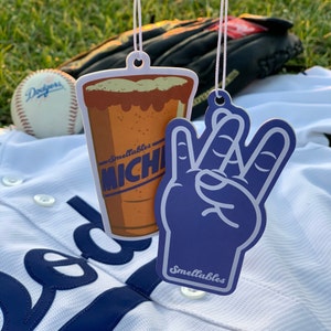 Puede incluir: Dos ambientadores de coche colgados de una camiseta de béisbol blanca. Un ambientador tiene forma de jarra de cerveza con el texto "Smelltables Michi" y el otro es una mano morada haciendo un signo de paz con el texto "Smelltables".
