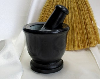 May include: A black granite mortar and pestle set. The mortar is a deep bowl with a rounded base and a smooth, polished surface. The pestle is a cylindrical shape with a rounded end.