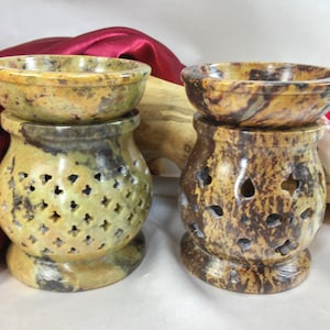 May include: Two stone oil warmers. The warmers have a bowl on top and a decorative base with cut-out designs. One is light green and the other is brown. The warmers are made of stone and are designed to hold essential oils.