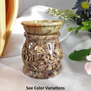 May include: A decorative stone oil warmer with a carved design. The warmer has a round bowl on top and a base with cut-out patterns. The color is a mix of beige, brown, and reddish hues. The text "See Color Variations" is at the bottom.