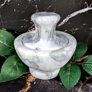 May include: A gray and white marble mortar and pestle set. The mortar is a bowl shape with a rounded base, and the pestle has a rounded handle. The set is displayed on a dark background with green leaves.