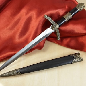 May include: A decorative dagger with a black handle and a silver blade. The dagger has intricate gold-colored metalwork on the handle and pommel. The dagger is lying on a red satin fabric.