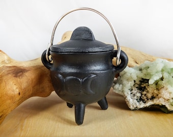 May include: A small black cast iron cauldron with a lid and a silver wire handle. The cauldron has three legs and a triple moon design on the side.