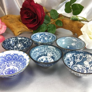 May include: Six small, blue and white patterned ceramic bowls with gold rims. The bowls feature various floral and abstract designs. The bowls are arranged on a light-colored surface, with flowers in the background.