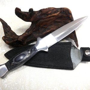 May include: A stainless steel dagger with a black and gray wooden handle rests in a black sheath. The dagger has a double-edged blade and a silver metal guard. A piece of driftwood is in the background.