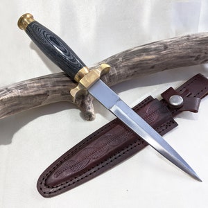 May include: A black and grey handled dagger with a brass pommel and a silver blade. The dagger is in a brown leather sheath with a snap closure.