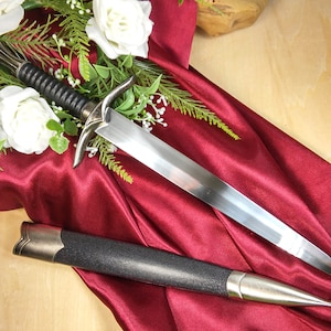 May include: A silver dagger with a black handle and a black leather scabbard. The dagger is lying on a red satin fabric with white roses.