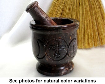 May include: A dark brown stone mortar and pestle with a triple moon design. The mortar is carved with a triple moon symbol and a face in the center.