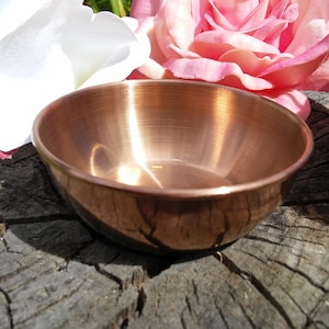 May include: A small, shiny copper bowl sits on a rough, wooden surface. The bowl is smooth and reflective, with a slight indentation in the center. The bowl is surrounded by pink and white flower petals.