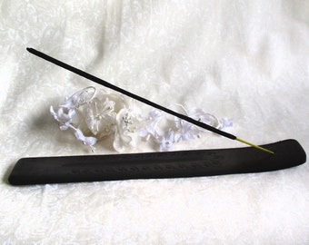 May include: A black incense stick holder with a carved design. The holder is made of wood and has a rectangular shape. A black incense stick is placed on the holder.