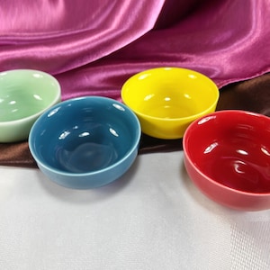 May include: Four ceramic bowls in different colors: green, blue, yellow, and red. The bowls are arranged in a semi-circle on a white surface.