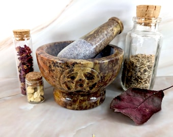 May include: A brown and green stone mortar and pestle with a carved pentagram design. The mortar and pestle are sitting on a white marble surface with three small glass jars with cork tops filled with dried herbs and flowers.