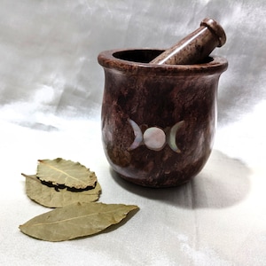 May include: A brown stone mortar and pestle with a triple moon design made of mother of pearl. The mortar and pestle are sitting on a white surface with three bay leaves.
