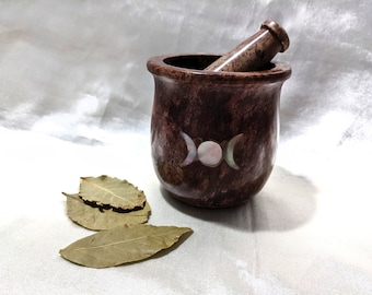 May include: A brown stone mortar and pestle with a triple moon design made of mother of pearl. The mortar and pestle are sitting on a white surface with three bay leaves.