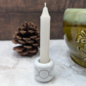 May include: A white candle in a white ceramic holder featuring a triple moon design. The candle is central, with a pinecone and a green ceramic mug in the background. The candle holder is on a marble surface.