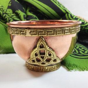 May include: A copper bowl with a brass trim and a Celtic knot design. The bowl is sitting on a green and black fabric.