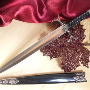 May include: A silver and black dagger with a skull design on the handle and a black leather sheath. The dagger is lying on a wooden surface with a red sparkly leaf.