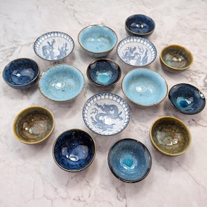 May include: An assortment of small ceramic bowls in shades of blue, turquoise, and brown. Some bowls feature a dragon design in white and blue. The bowls are arranged on a marble surface.