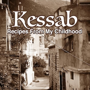 Op de afbeelding: Een zwart-witfoto van een smalle straat in een dorp. De straat is omzoomd met stenen gebouwen en er loopt een persoon over straat. De tekst "Kessab Recipes From My Childhood by Zevart Keshishian" staat onderaan de afbeelding.