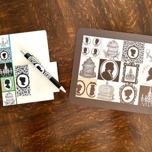 May include: A set of note cards with a black and white silhouette design featuring a birdcage, chandelier, and a person's profile. The note cards are on a brown wooden surface with a pen.