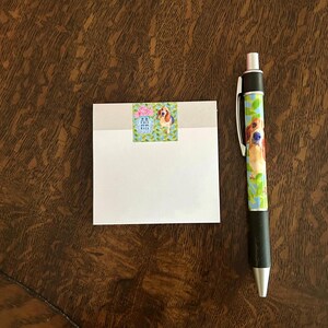 May include: A white notepad with a sticker of a dog on a blue and green background. A pen with a dog design is next to the notepad on a brown wooden surface.