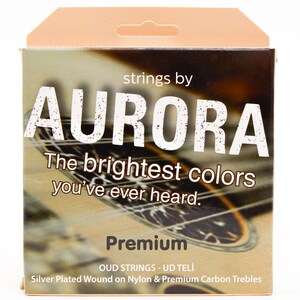 Könnte beinhalten: Eine Schachtel Oud-Saiten der Marke Aurora. Die Schachtel ist beige und braun mit den Worten "AURORA" und "The brightest colors you've ever heard." auf der Vorderseite. Auf der Schachtel steht auch "Premium" und die Saitenmaterialien sind angegeben.