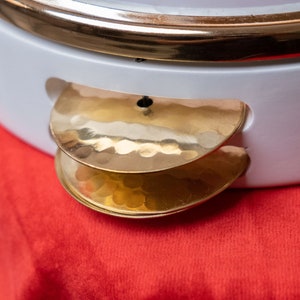 May include: A white tambourine with two gold cymbals attached to the side. The cymbals are hammered and have a textured surface.