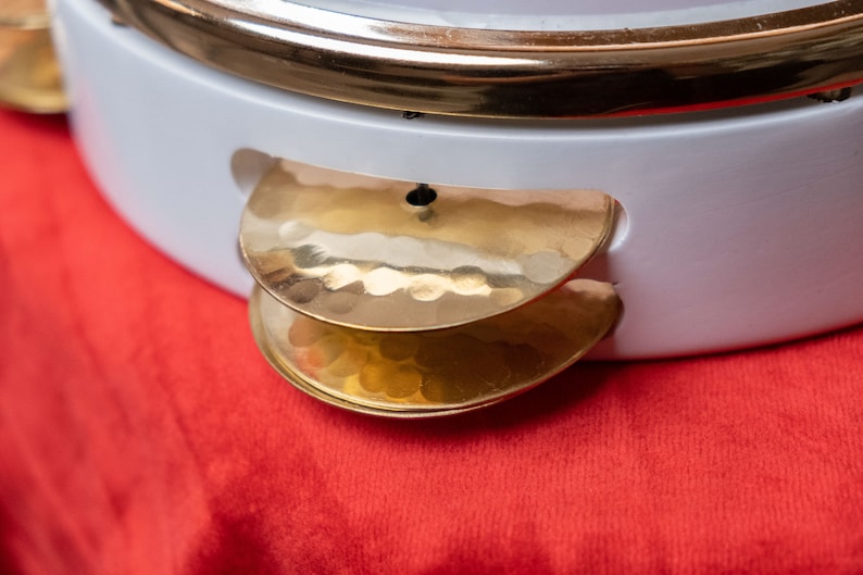 May include: A white tambourine with two gold cymbals attached to the side. The cymbals are hammered and have a textured surface.